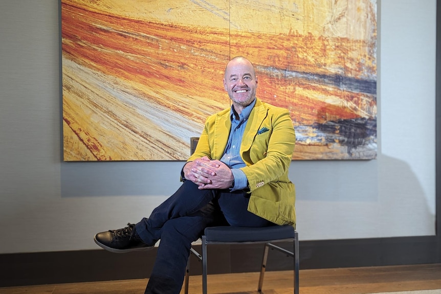 Adam wears a yellow jacket and blue pants and sits in front of a yellow and orange hued painting