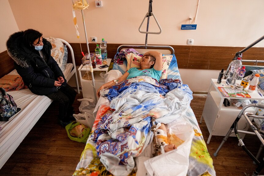 A woman lies in a hospital bed with an injured leg, speaking to a woman sitting on another bed next to her.