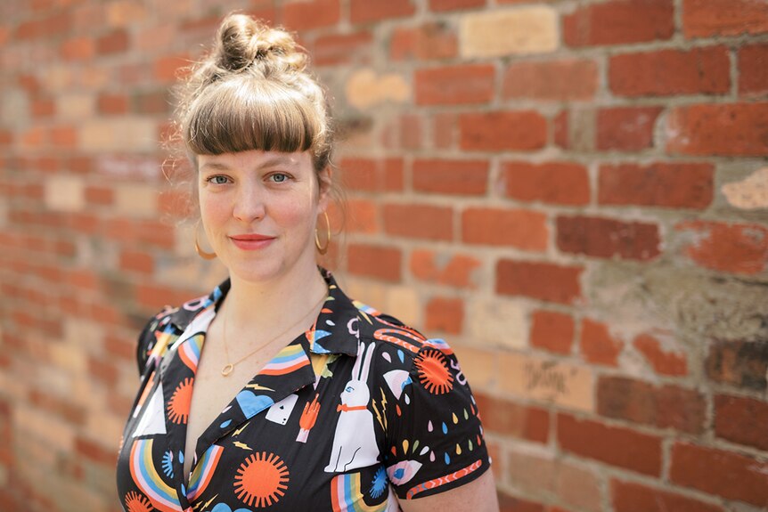 Sarah Firth wears shirt decorated with bright graphics and stands in front of red brick wall outdoors, hair glistens from sun.