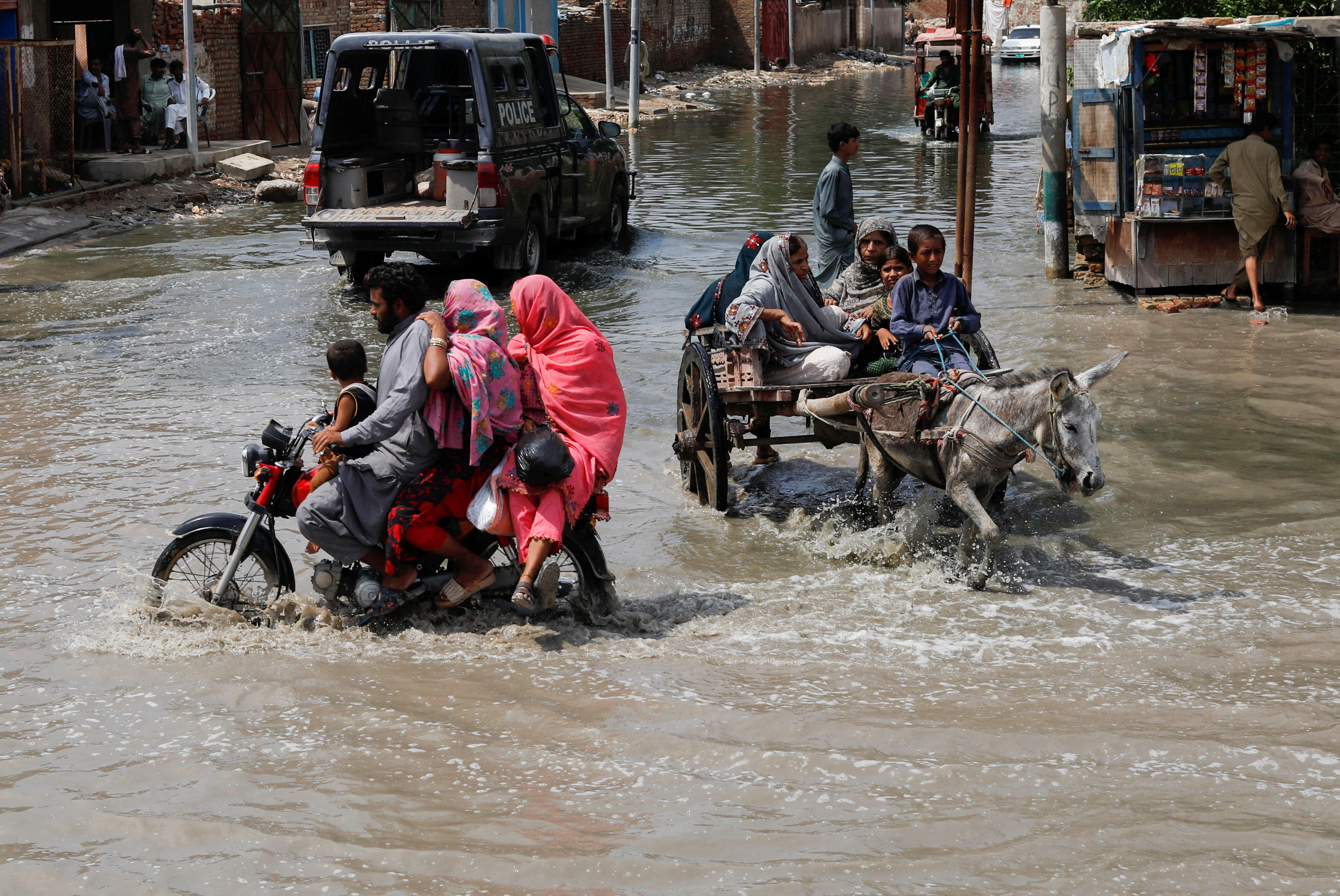 a family on a motorbike and people on a cart drawn by a donkey travel down a street in knee deep water.
