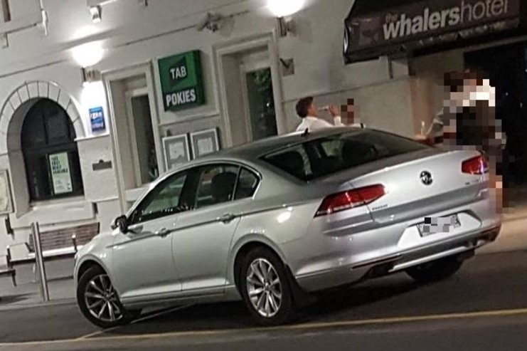 A man stands next two a car and has a beer, unaware that his photo is being taken.