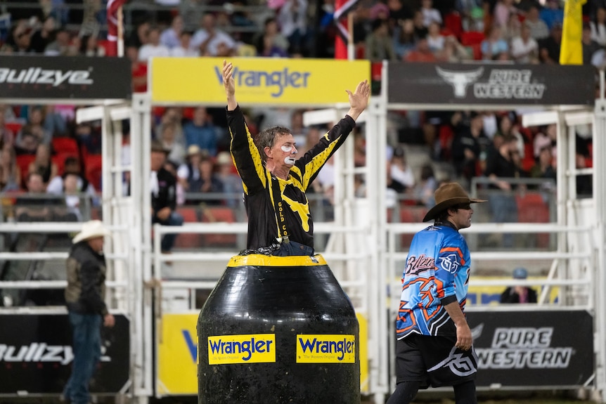 Man dressed in clown costume and makeup appearing from barrel in rodeo ring. 