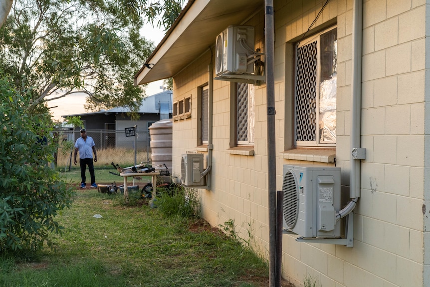 A man stands outside a home that has three air conditioning units attached to a wall.