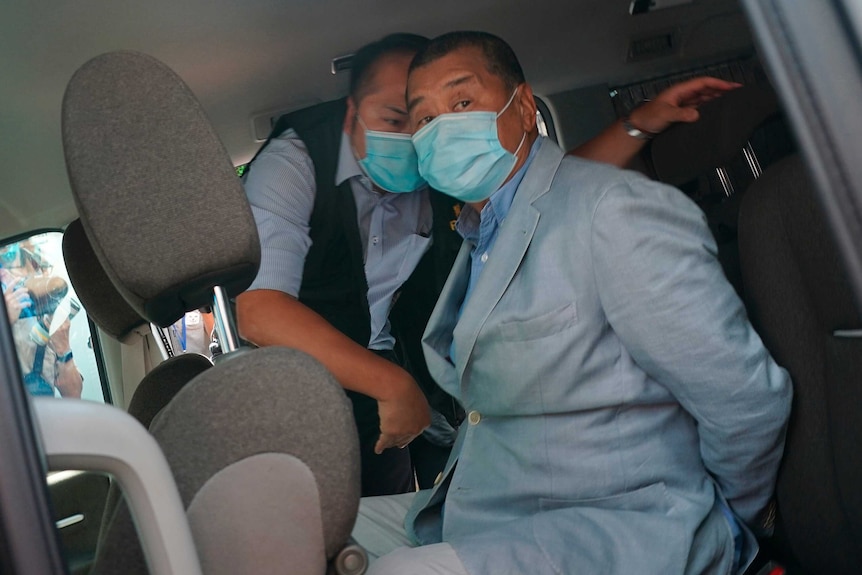 A man in a suit and face mask with his hands behind his back in the back seat of a car.