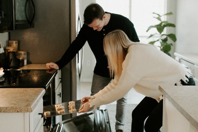 A man watches as his partner puts a tray of goods into an oven.