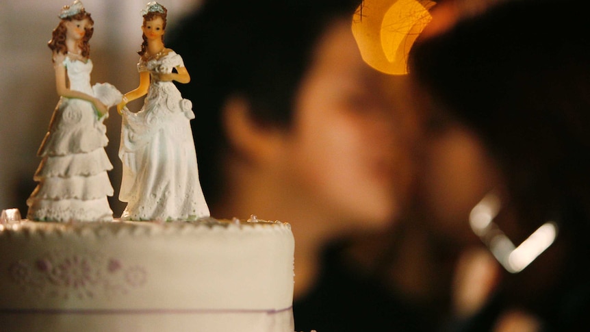 Two female figurines on a top of a cake with two women kissing behind the cake.