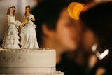 Two female figurines on a top of a cake with two women kissing behind the cake.