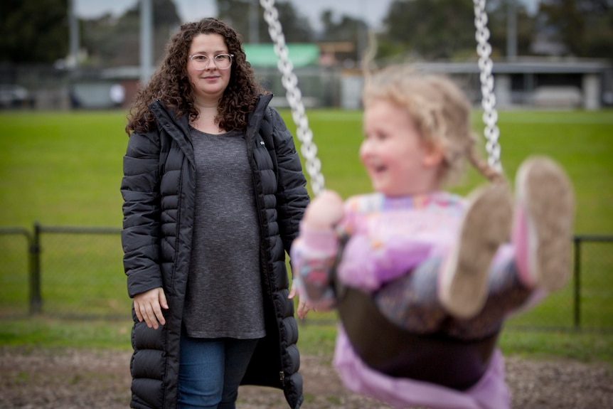 A young white woman watching on as a toddler plays on a swing