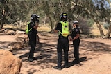 Three police officers stand near each other on red dirt near gum trees