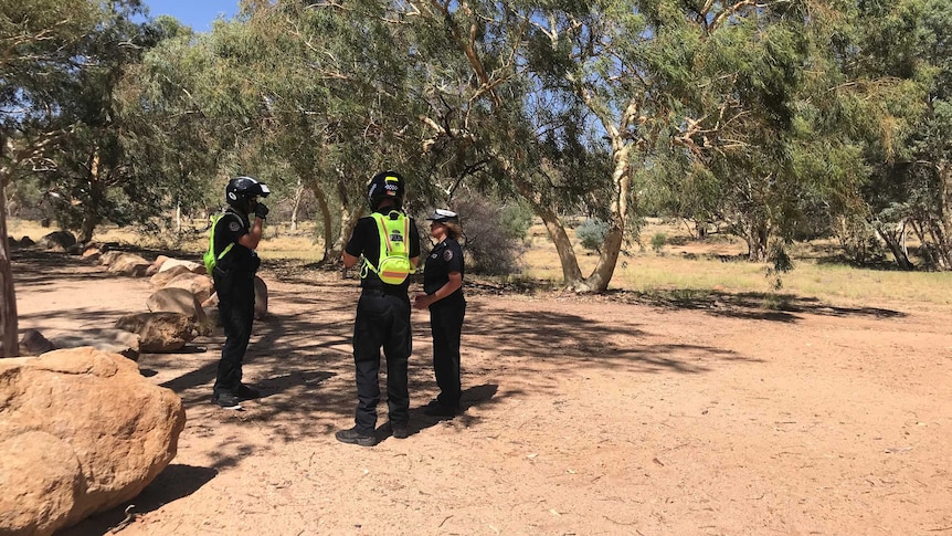 Three police officers stand near each other on red dirt near gum trees