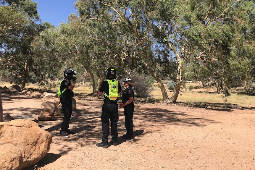 Three police officers stand near each other on red dirt near gum trees