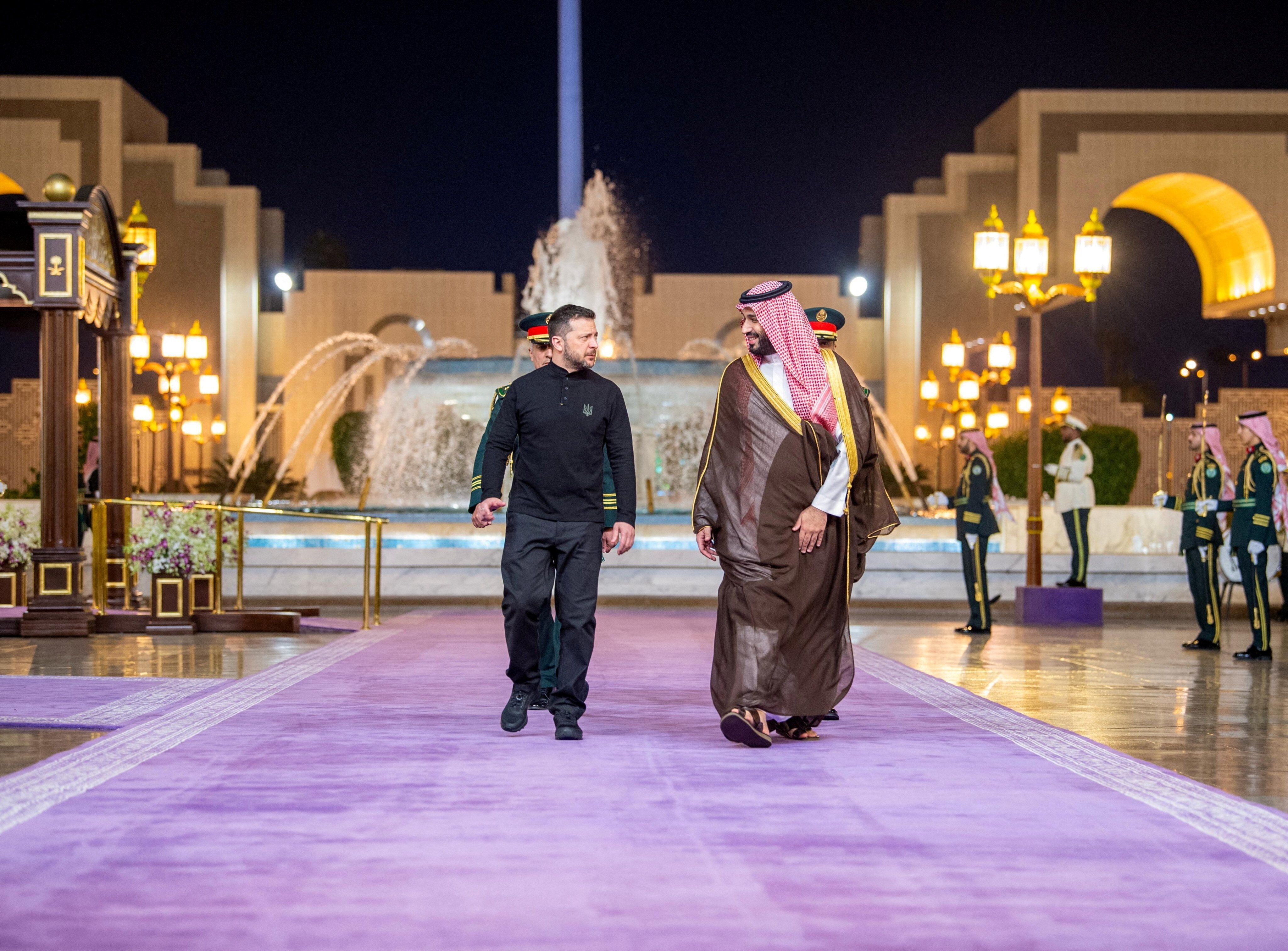 A man in a black outfit walks with a man in a Saudi thwab