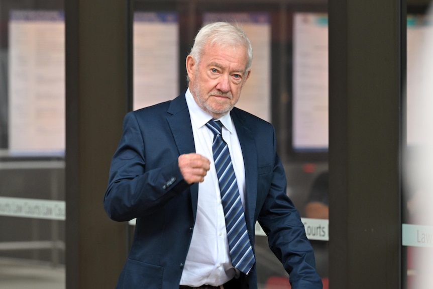 A white haired man in a suit walks outside a court building.