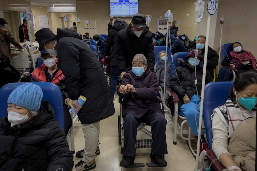 A man pushes an elderly person in a wheelchair through a crowded hospital corridor