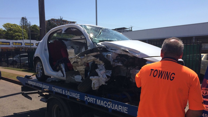 A white car that has been involved in a crash, on the back of a truck, with a ma in an orange shirt on the right.