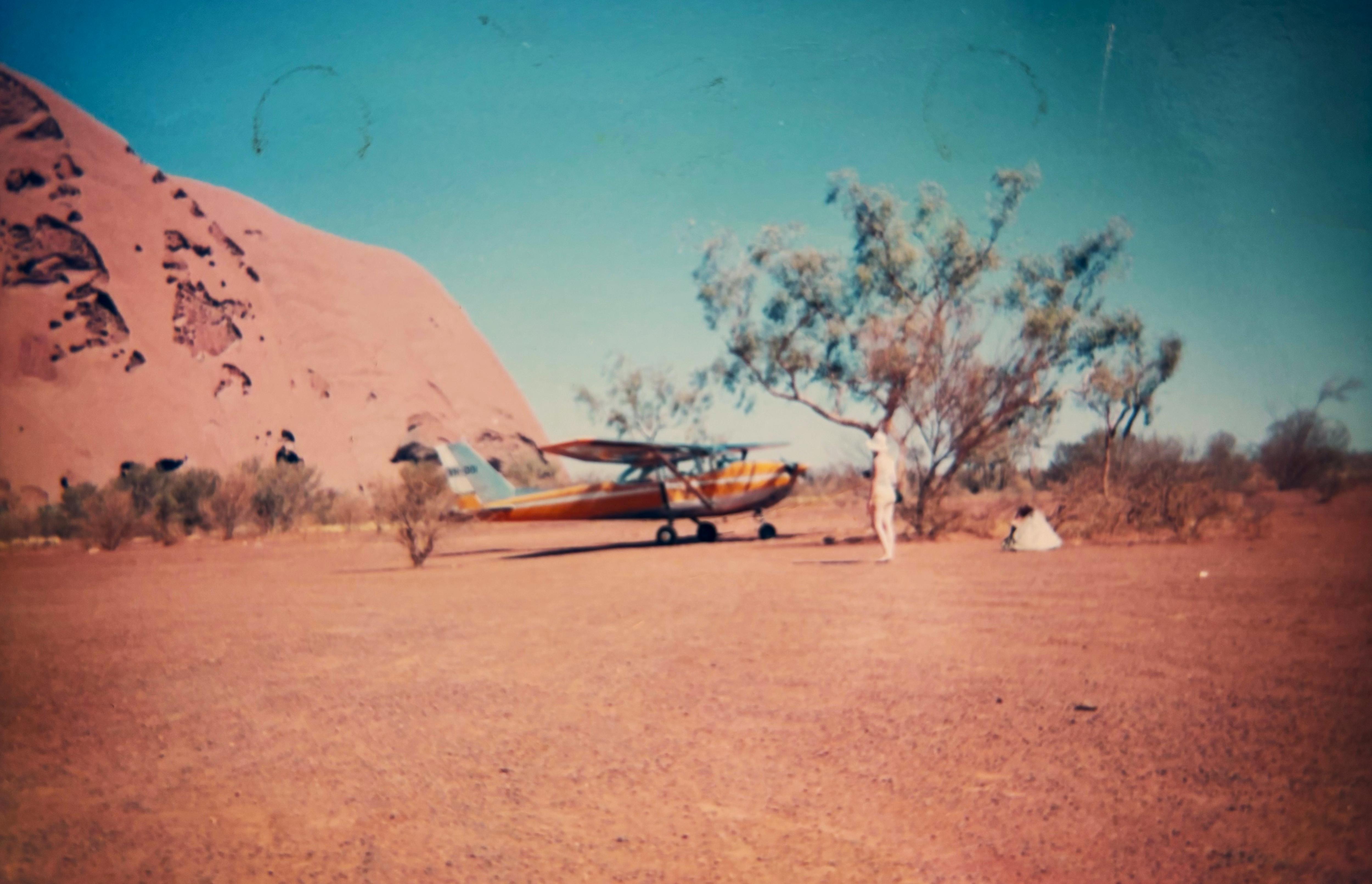 Part of Uluru, red dusty rock, on left of photo with small yellow and white plane near tree on right.
