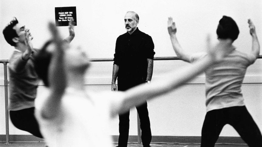 In a dance studio, Jerome Robbins stands by the wall watching three dancers move around him with their arms raised.