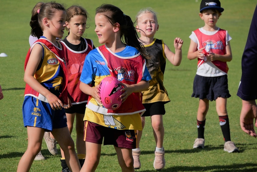 A group of girls run around wearing football shirts and holding a pink ball.
