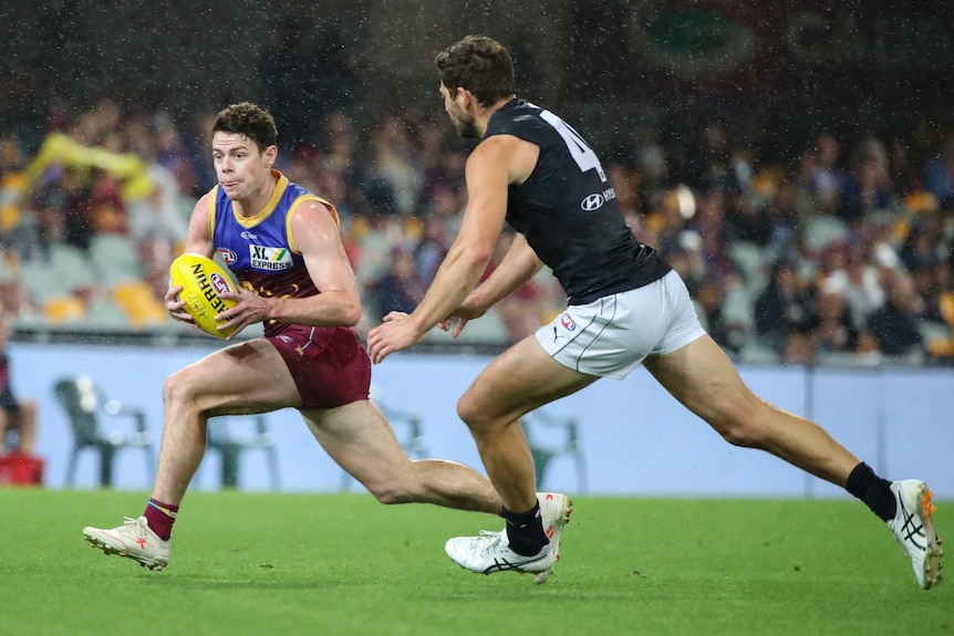 A Brisbane Lions AFL player holds the ball in both hands as he tries to run around a Carlton opponent.