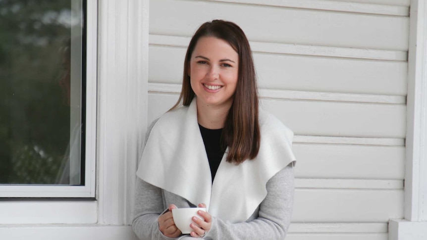 Caucasian woman with brown hair wearing a grey jacket sits on a bench against a white house holding a tea cup, smiling.