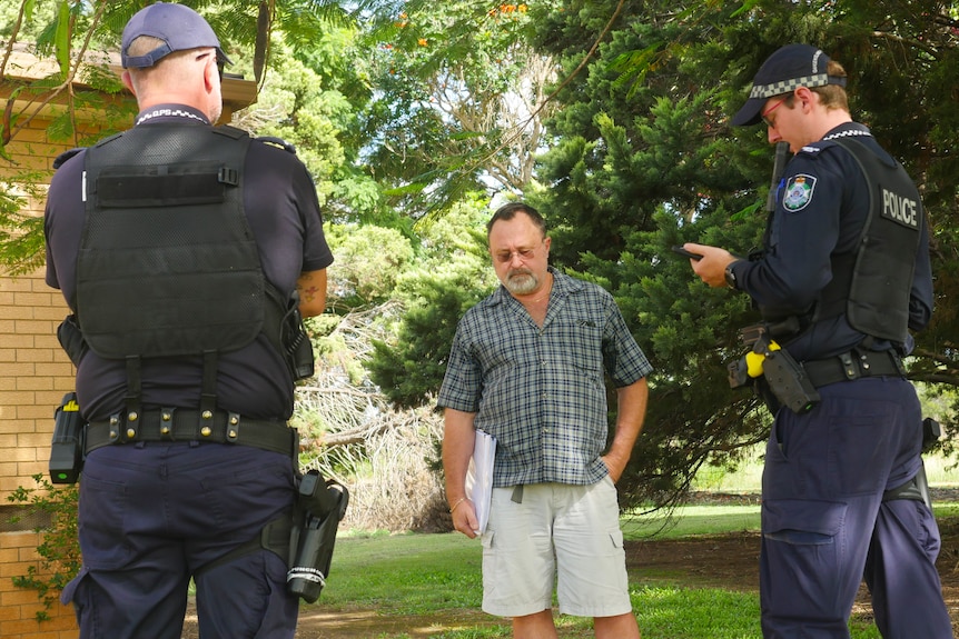 A wide shot of a man facing the camera flanked by police who face away.