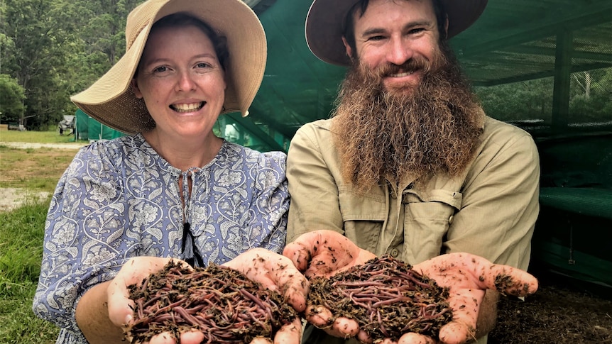 A lady in a floppy hat, and a  bloke with a big bushy beard and hat hold up earthworms in their hands.