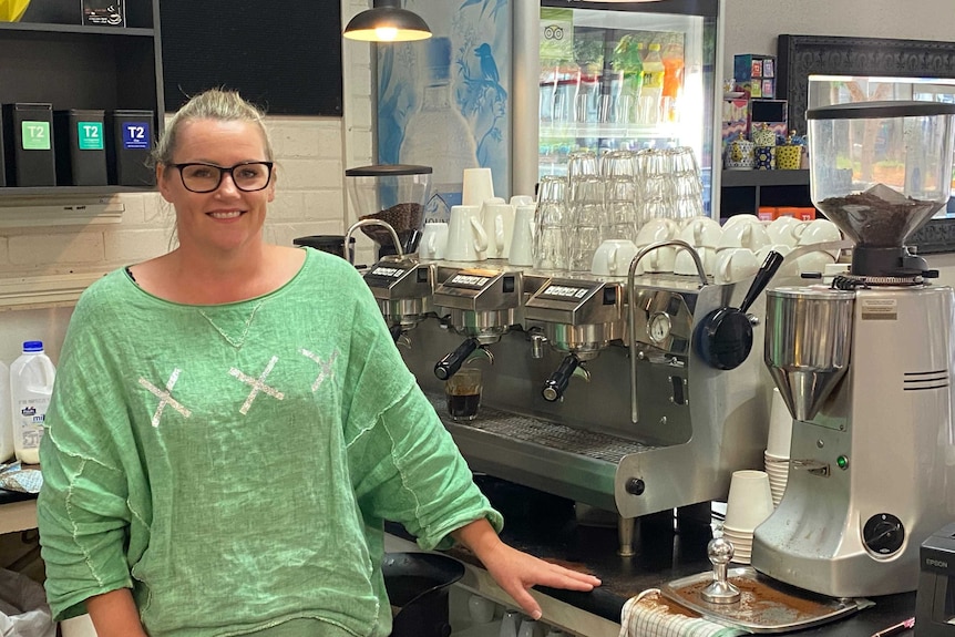 A woman in a green blouse stands in front of a coffee machine in an artificially lit room.