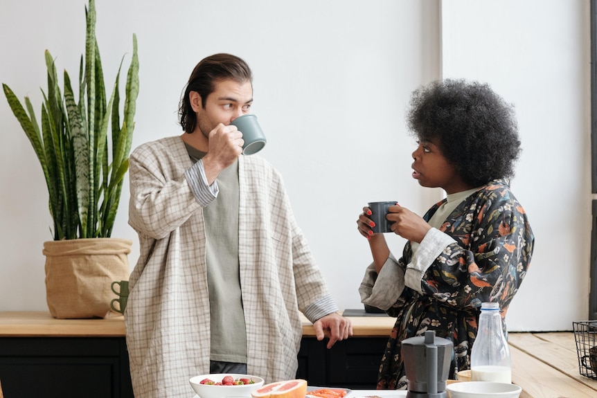 A couple stand together in a kitchen drinking coffee. The man and woman look at each other with watchful eyes.  
