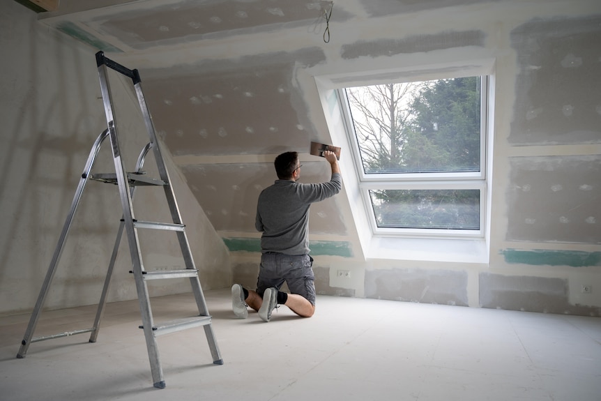 A man kneels on the floor plastering drywall next to a window.