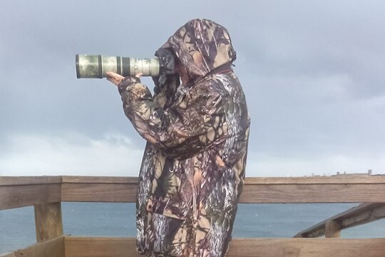 A person wearing camouflage poses on a sea deck with a long lensed camera
