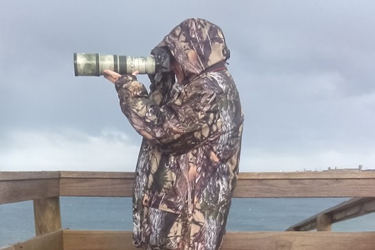 A person wearing camouflage poses on a sea deck with a long lensed camera