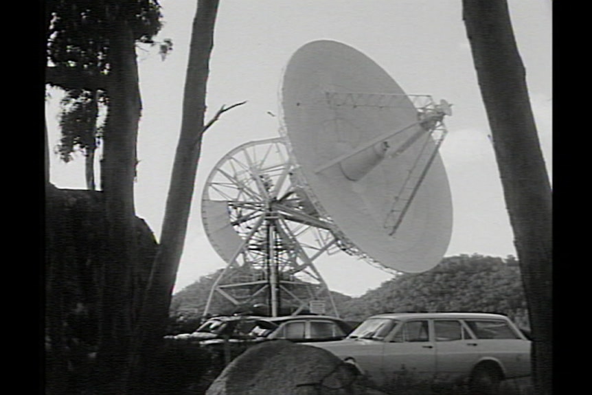 The Honeysuckle Creek satellite is seen in black and white, some cars in the foreground.