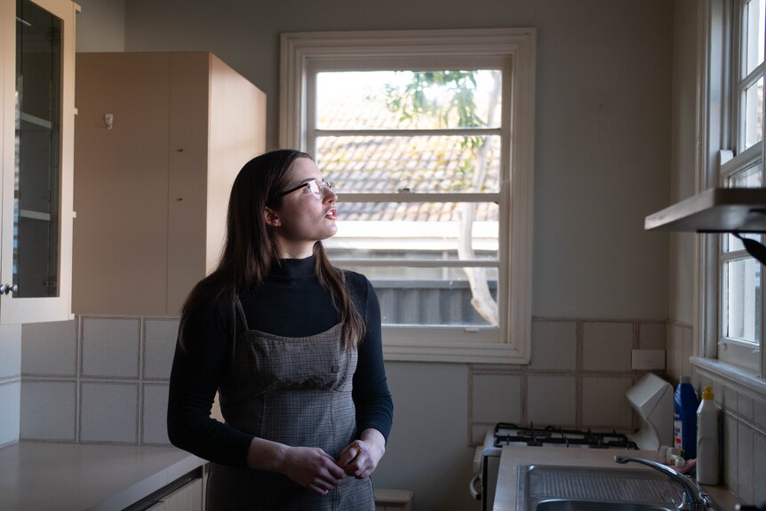 A woman wearing black skivvy under grey pinafore dress stands in a kitchen, looking to window