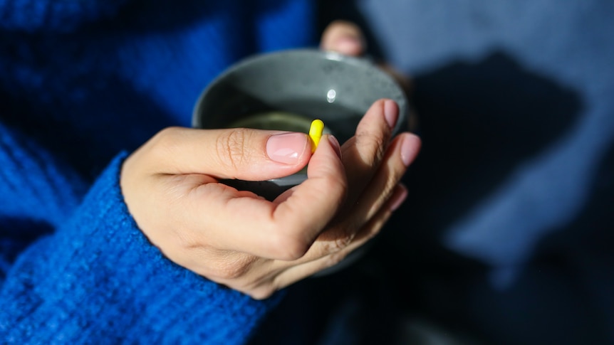 Close-up shows woman's hand in blue jumper sleeve holding a yellow pill between two fingers while clutching a mug of lemon water