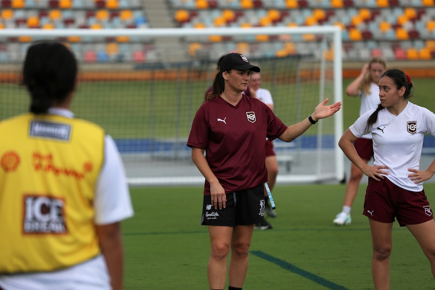 A woman is gesturing to players. She is coaching them. She wears a burgundy shirt. The players around her listen carefully.