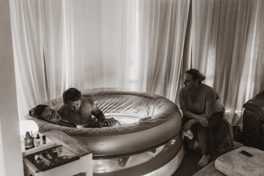 A woman leans back as she sits in a shallow pool inside her home and gives birth with her husband in the water with her.