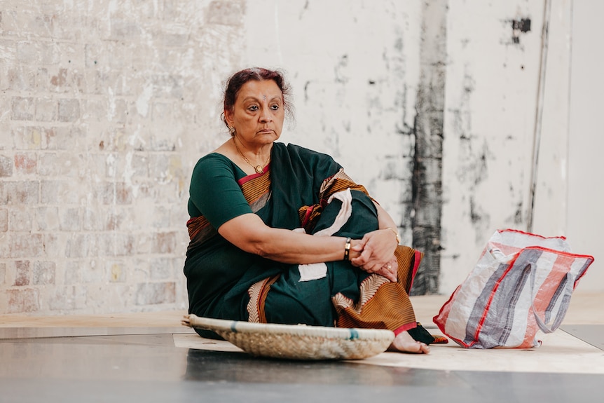 A Sri Lankan woman in her mid 60s in a green sari, sits on a stage, arms clasped, looking concerned