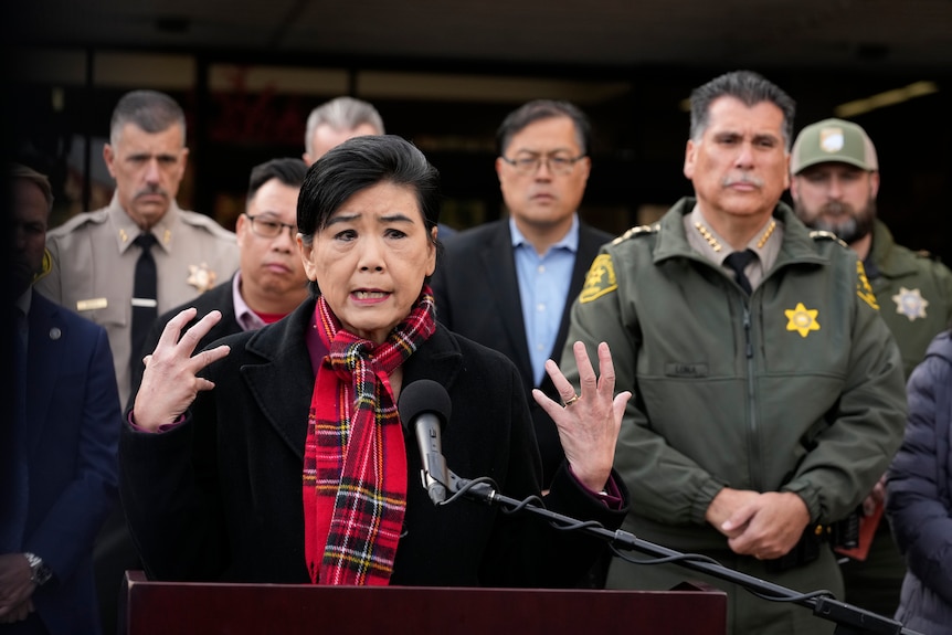 An Asian woman speaks into a microphone as people in suits and police uniforms stand behind her. 