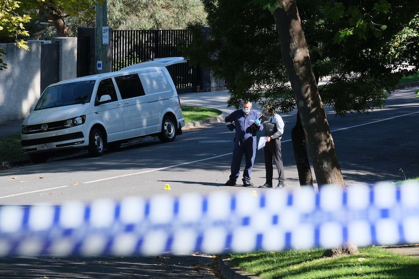 Detectives stand behind police tape on a leafy street.