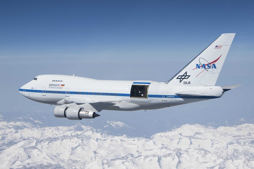 A shorter, stockier than usual 747 flies above the clouds with a large door open towards the rear of the aircraft.