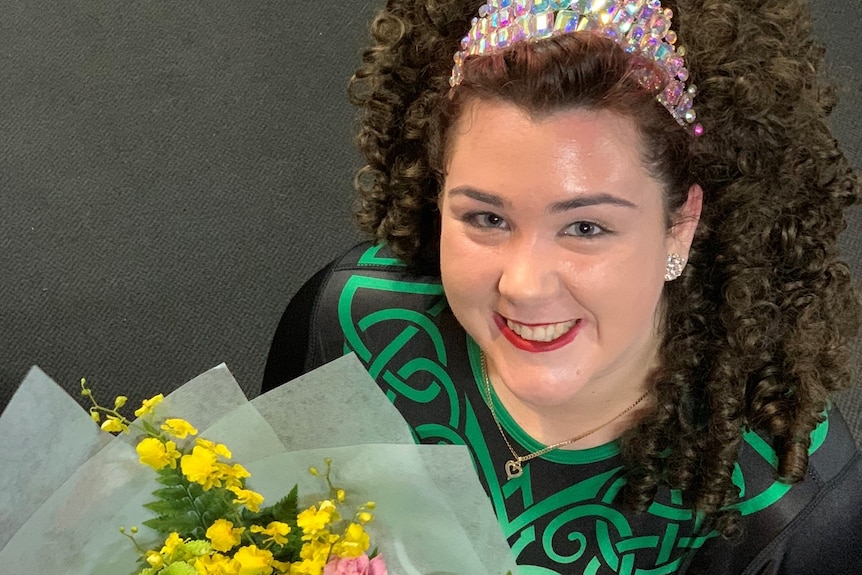 A young girl with curly brown hair wearing a black and green dance costume holds a bright bouquet of flowers