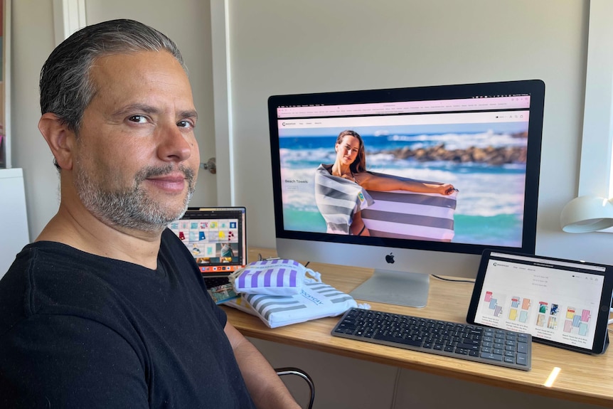 A man wearing a black t-shirt sits at a computer showing a woman wrapped in a towel on a beach.