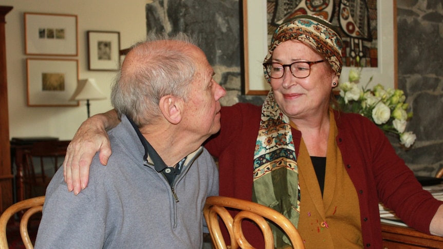 A woman with a hair wrap has her arm around a man as they sit on chairs in a home.