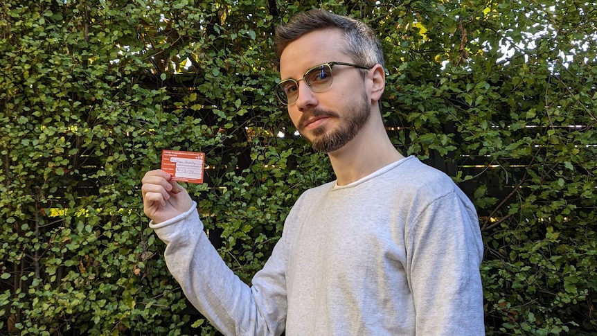 A man holds up a vaccination card in front of a hedge, smiling.