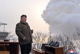 Kim Jong Un with a cigarette in his hand, smiling as a big cloud of smoke rises behind him.