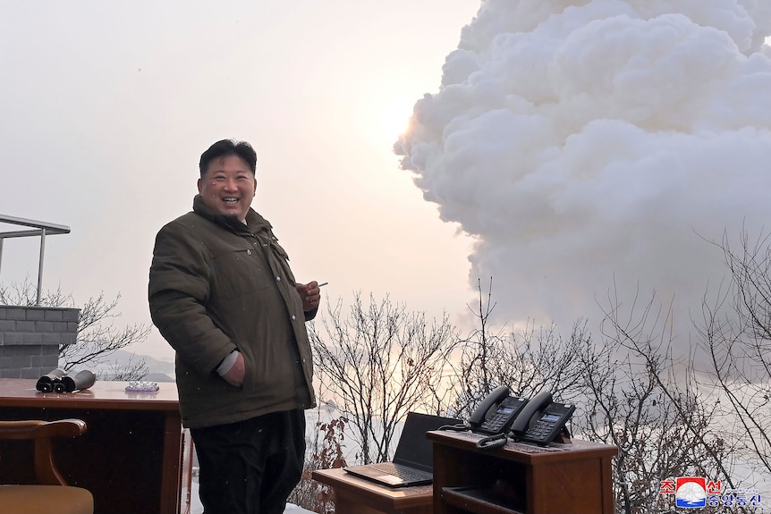 Kim Jong Un with a cigarette in his hand, smiling as a big cloud of smoke rises behind him.
