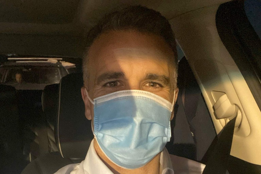 A man in a white shirt sitting in his car wearing a face mask.
