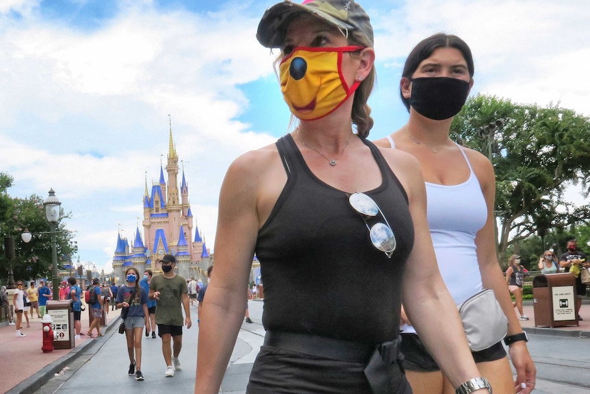 Two young women in summer clothing wear face masks as they walk along a footpath at amusement park.