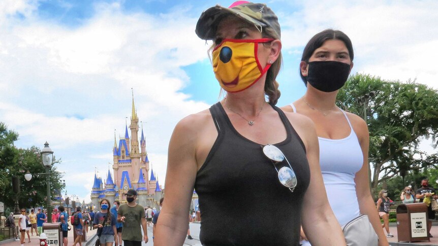 Two young women in summer clothing wear face masks as they walk along a footpath at amusement park.