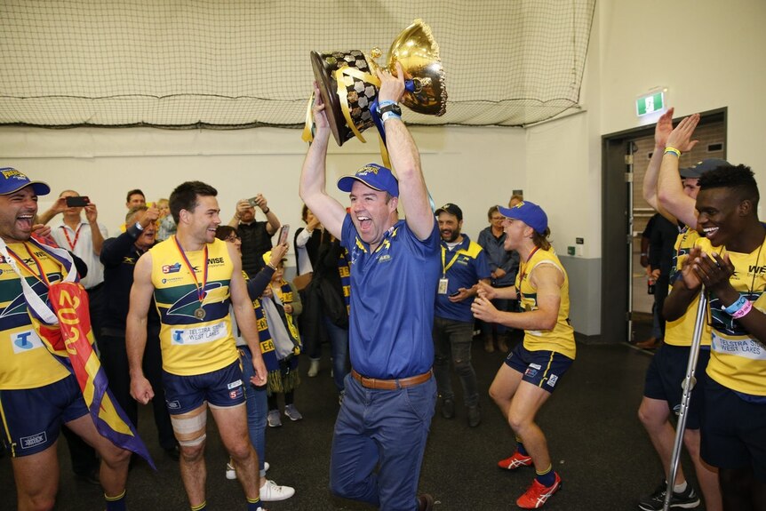 A man holds a large gold cup aloft while surrounded by football players.